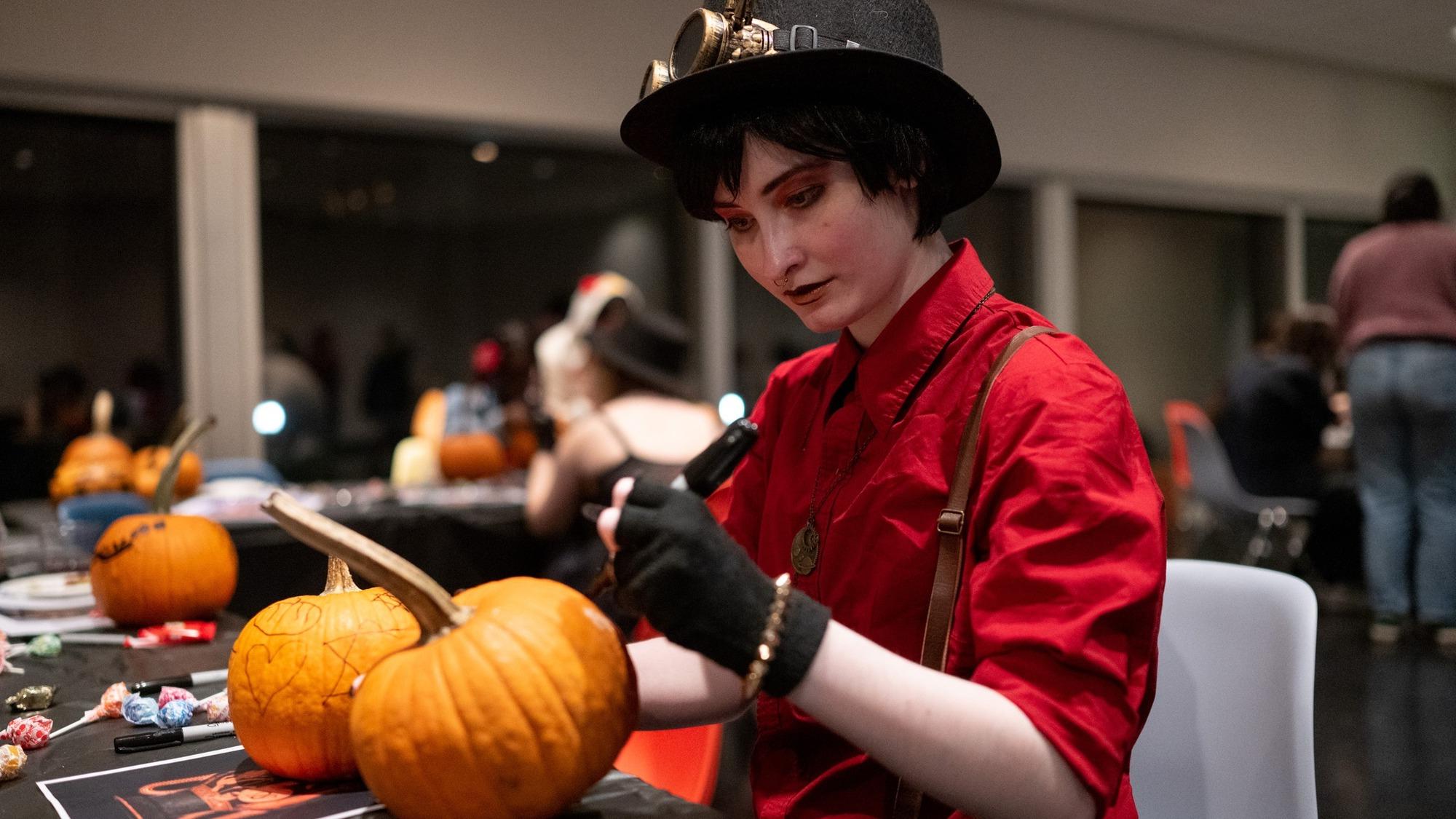 A student in an outfit that evokes the steampunk aesthetic decorates pumpkins at a table in the Tang.